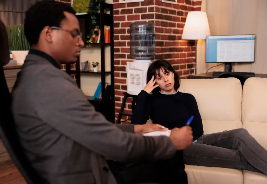 Male psychiatrist in a grey suit holding a pen and notepad, seated across from a female patient who rests her hand on her forehead with a worried expression while sitting on a cream sofa in an office with an exposed brick wall, bookshelf, and computer desk.